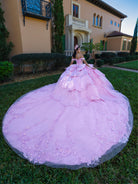 This gown features a fitted, embellished bodice with off-the-shoulder sleeves and a full tiered ball-gown skirt. Intricate appliqués decorate the layers, and a long flowing train with a large back bow completes the princess-style look.