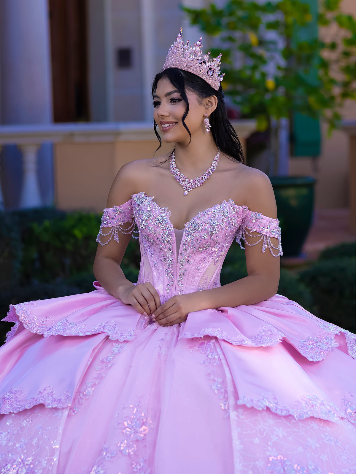 This gown features a fitted, embellished bodice with off-the-shoulder sleeves and a full tiered ball-gown skirt. Intricate appliqués decorate the layers, and a long flowing train with a large back bow completes the princess-style look.