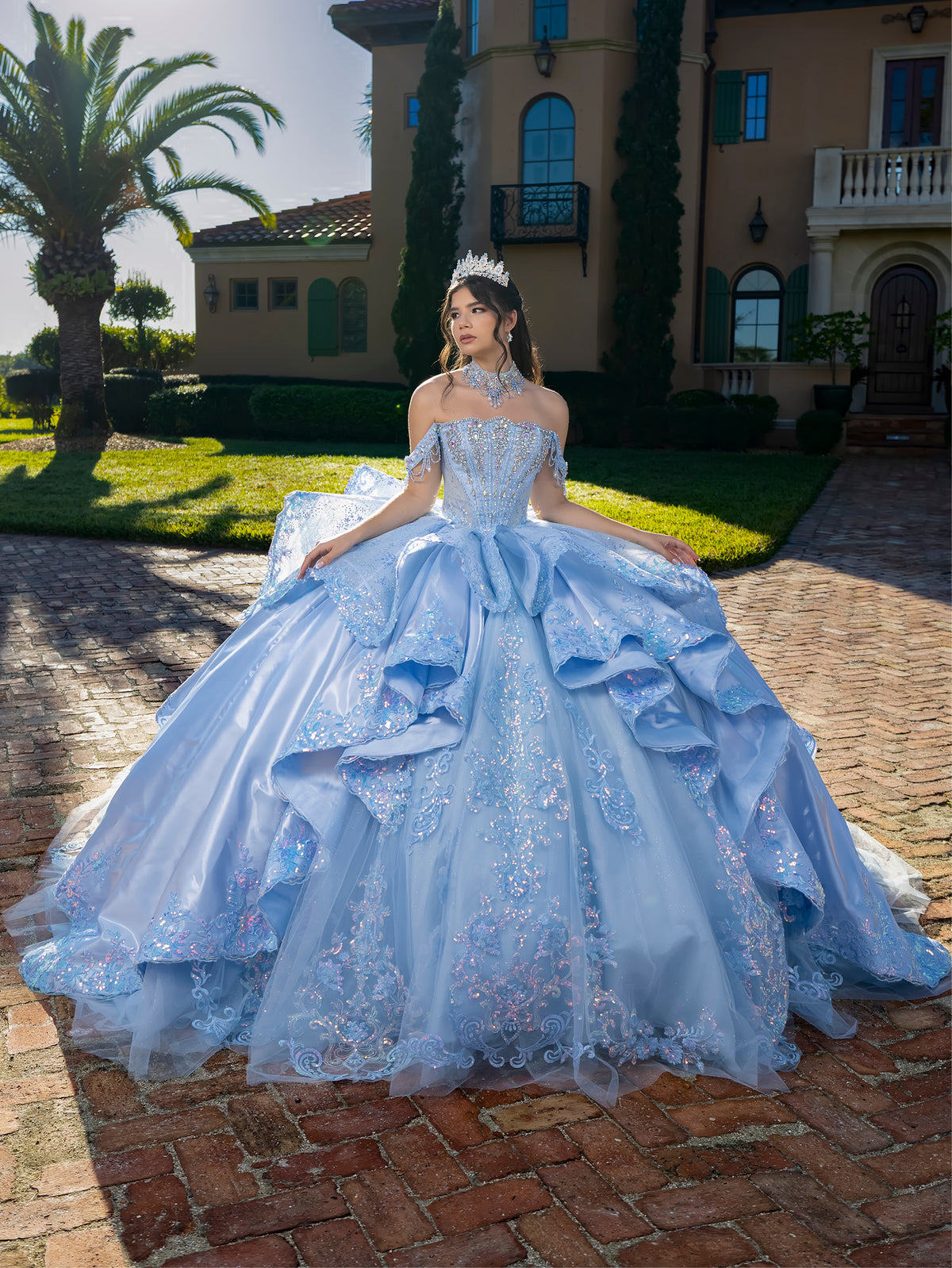 Woman in a light blue gown sitting on a brick path with a mansion in the background / This gown features a detailed off-the-shoulder bodice and a full layered ball-gown skirt. A large, sculpted bow highlights the back, leading into a long train decorated with ornate appliqués for a dramatic, regal finish.
