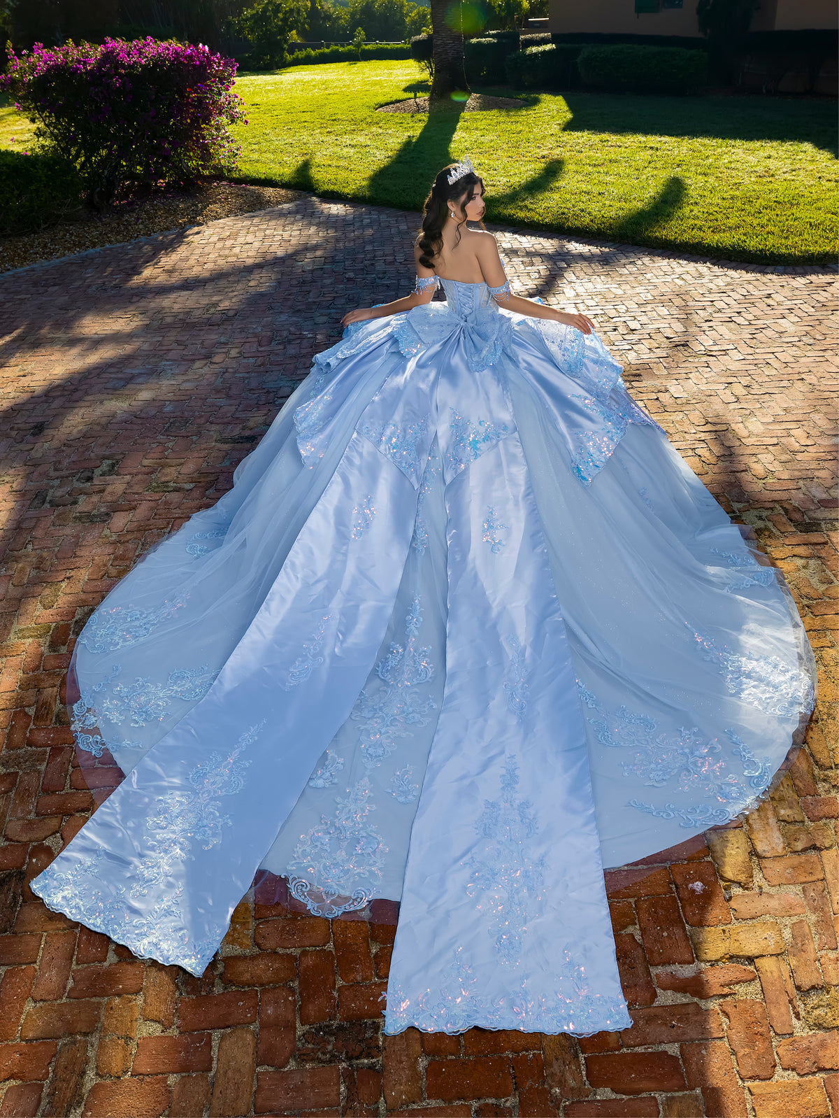 Woman in a light blue wedding dress standing on a brick path with greenery in the background / This gown features a detailed off-the-shoulder bodice and a full layered ball-gown skirt. A large, sculpted bow highlights the back, leading into a long train decorated with ornate appliqués for a dramatic, regal finish.