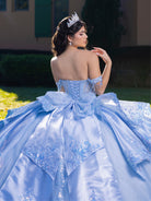 A woman wearing a voluminous light blue ball gown with lace appliqué, a corset-style back, and a large bow at the waist, photographed outdoors on grass with a building in the background. She is seen from the back and wears a tiara. / This gown features a detailed off-the-shoulder bodice and a full layered ball-gown skirt. A large, sculpted bow highlights the back, leading into a long train decorated with ornate appliqués for a dramatic, regal finish.