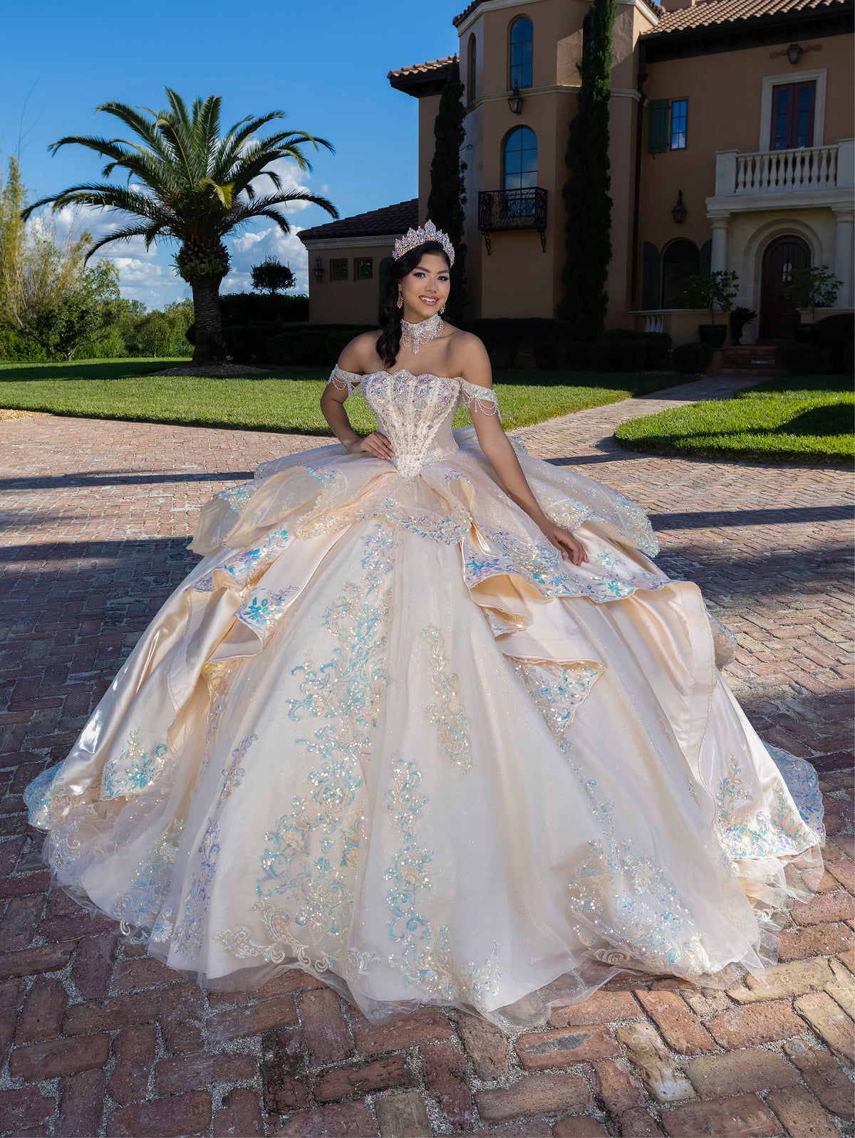 Woman in an elegant white gown with floral details standing in front of a mansion. / This gown features a detailed off-the-shoulder bodice and a full layered ball-gown skirt. A large, sculpted bow highlights the back, leading into a long train decorated with ornate appliqués for a dramatic, regal finish.