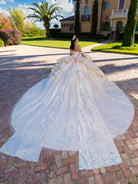 White wedding dress with a long train on a brick driveway in front of a large house. / This gown features a detailed off-the-shoulder bodice and a full layered ball-gown skirt. A large, sculpted bow highlights the back, leading into a long train decorated with ornate appliqués for a dramatic, regal finish.