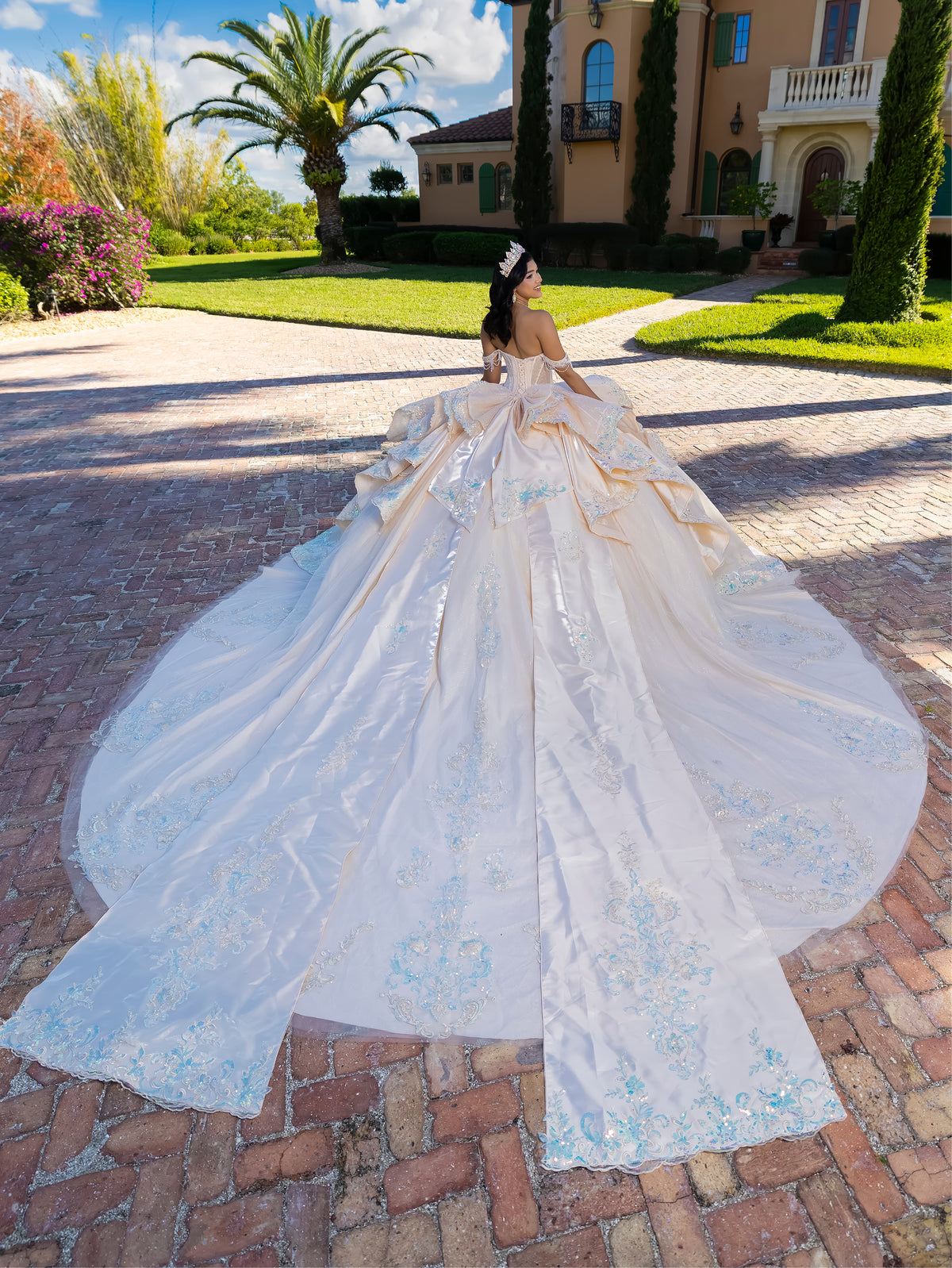 White wedding dress with a long train on a brick driveway in front of a large house. / This gown features a detailed off-the-shoulder bodice and a full layered ball-gown skirt. A large, sculpted bow highlights the back, leading into a long train decorated with ornate appliqués for a dramatic, regal finish.