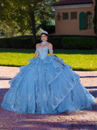 Woman in a blue quinceañera dress standing outdoors on a brick path with greenery and a building in the background. / This gown features a fitted, embellished bodice with off-the-shoulder straps and a full layered ball-gown skirt. Intricate appliqués decorate the skirt, while a large, sculpted bow and long ornate train complete the dramatic, regal design.