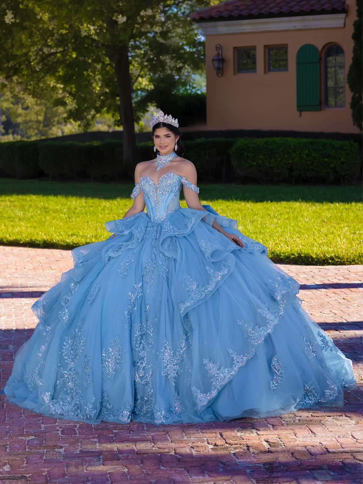 Woman in a blue quinceañera dress standing outdoors on a brick path with greenery and a building in the background. / This gown features a fitted, embellished bodice with off-the-shoulder straps and a full layered ball-gown skirt. Intricate appliqués decorate the skirt, while a large, sculpted bow and long ornate train complete the dramatic, regal design.
