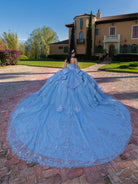 A woman wearing a voluminous light blue ball gown with intricate lace embroidery stands on a brick courtyard in front of a large yellow villa. / This gown features a fitted, embellished bodice with off-the-shoulder straps and a full layered ball-gown skirt. Intricate appliqués decorate the skirt, while a large, sculpted bow and long ornate train complete the dramatic, regal design.