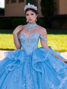 Woman in a blue quinceañera dress with intricate details and a tiara, standing outdoors. / This gown features a fitted, embellished bodice with off-the-shoulder straps and a full layered ball-gown skirt. Intricate appliqués decorate the skirt, while a large, sculpted bow and long ornate train complete the dramatic, regal design.