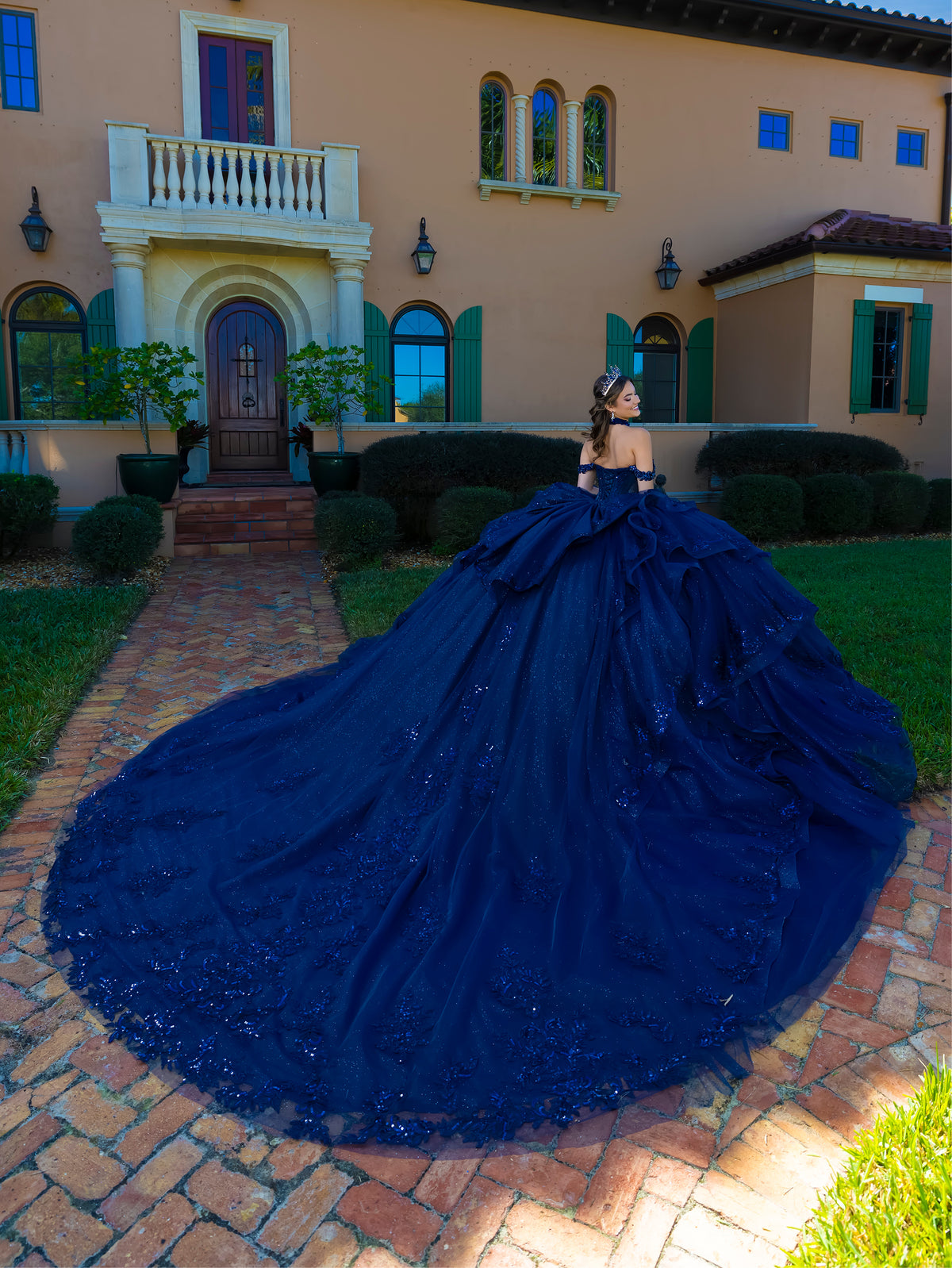 A woman wearing an elaborate off-the-shoulder royal blue ball gown with a massive train stands on a brick path in front of a large beige villa with green shutters. / This gown features a fitted, embellished bodice with off-the-shoulder straps and a full layered ball-gown skirt. Intricate appliqués decorate the skirt, while a large, sculpted bow and long ornate train complete the dramatic, regal design.