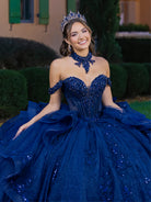 Woman in a blue sequined gown with a tiara outdoors. / This gown features a fitted, embellished bodice with off-the-shoulder straps and a full layered ball-gown skirt. Intricate appliqués decorate the skirt, while a large, sculpted bow and long ornate train complete the dramatic, regal design.