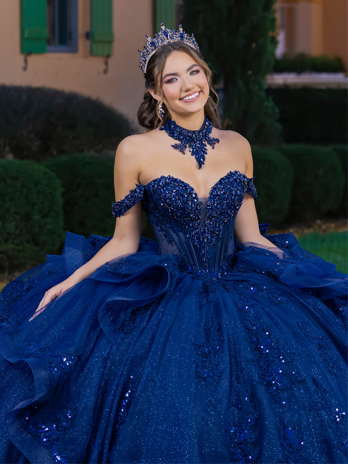 Woman in a blue sequined gown with a tiara outdoors. / This gown features a fitted, embellished bodice with off-the-shoulder straps and a full layered ball-gown skirt. Intricate appliqués decorate the skirt, while a large, sculpted bow and long ornate train complete the dramatic, regal design.