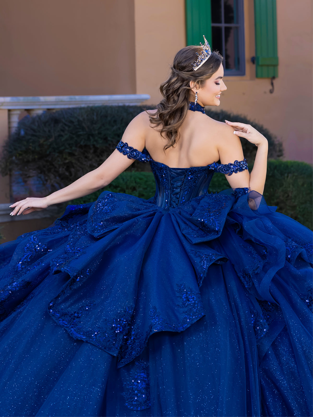 A woman wearing an elaborate royal blue off-the-shoulder ball gown with a corseted back, sheer sleeves, and a voluminous layered skirt with sequined details, posed outdoors in front of a building with green shutters. / This gown features a fitted, embellished bodice with off-the-shoulder straps and a full layered ball-gown skirt. Intricate appliqués decorate the skirt, while a large, sculpted bow and long ornate train complete the dramatic, regal design.