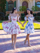 Two women in matching floral off-the-shoulder mini dresses holding hands while walking on a brick path in front of a house. One dress has a pink floral pattern on white, and the other has a blue floral pattern on white. / This gown features a full ball-gown skirt with delicate floral detailing and a fitted bodice with off-the-shoulder sleeves. A long, flowing train completes the elegant, fairytale look.