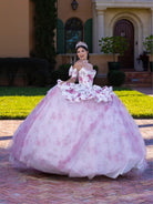 A woman wearing a voluminous off-the-shoulder quinceañera dress in light pink and white with a floral pattern, standing outdoors in front of a house with green shutters and a brick pathway. / This gown features a full ball-gown skirt with delicate floral detailing and a fitted bodice with off-the-shoulder sleeves. A long, flowing train completes the elegant, fairytale look.