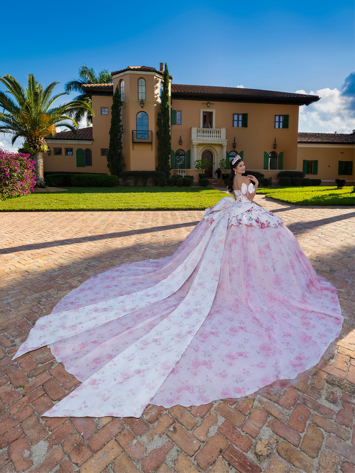 A woman wearing a voluminous light pink ball gown with a floral print and a long train stands on a brick patio in front of a large Mediterranean-style villa under a blue sky. / This gown features a full ball-gown skirt with delicate floral detailing and a fitted bodice with off-the-shoulder sleeves. A long, flowing train completes the elegant, fairytale look.