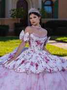 Woman in a floral dress with a tiara in an outdoor setting / This gown features a full ball-gown skirt with delicate floral detailing and a fitted bodice with off-the-shoulder sleeves. A long, flowing train completes the elegant, fairytale look.
