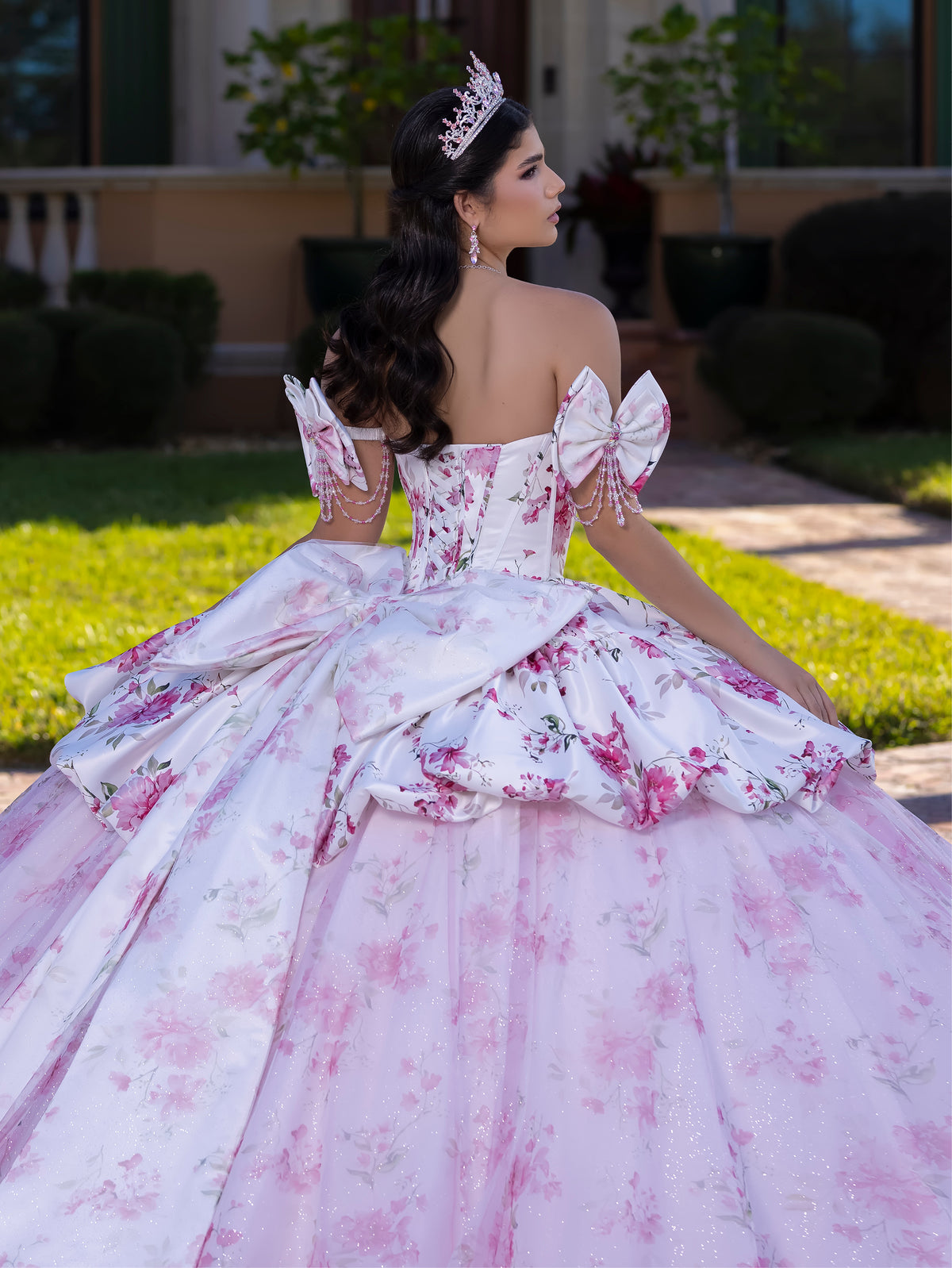 A woman wearing a formal off-the-shoulder ball gown with a pink and white floral pattern, photographed outdoors on a lawn with a house in the background. She is seen from behind, wearing a tiara. / This gown features a full ball-gown skirt with delicate floral detailing and a fitted bodice with off-the-shoulder sleeves. A long, flowing train completes the elegant, fairytale look.