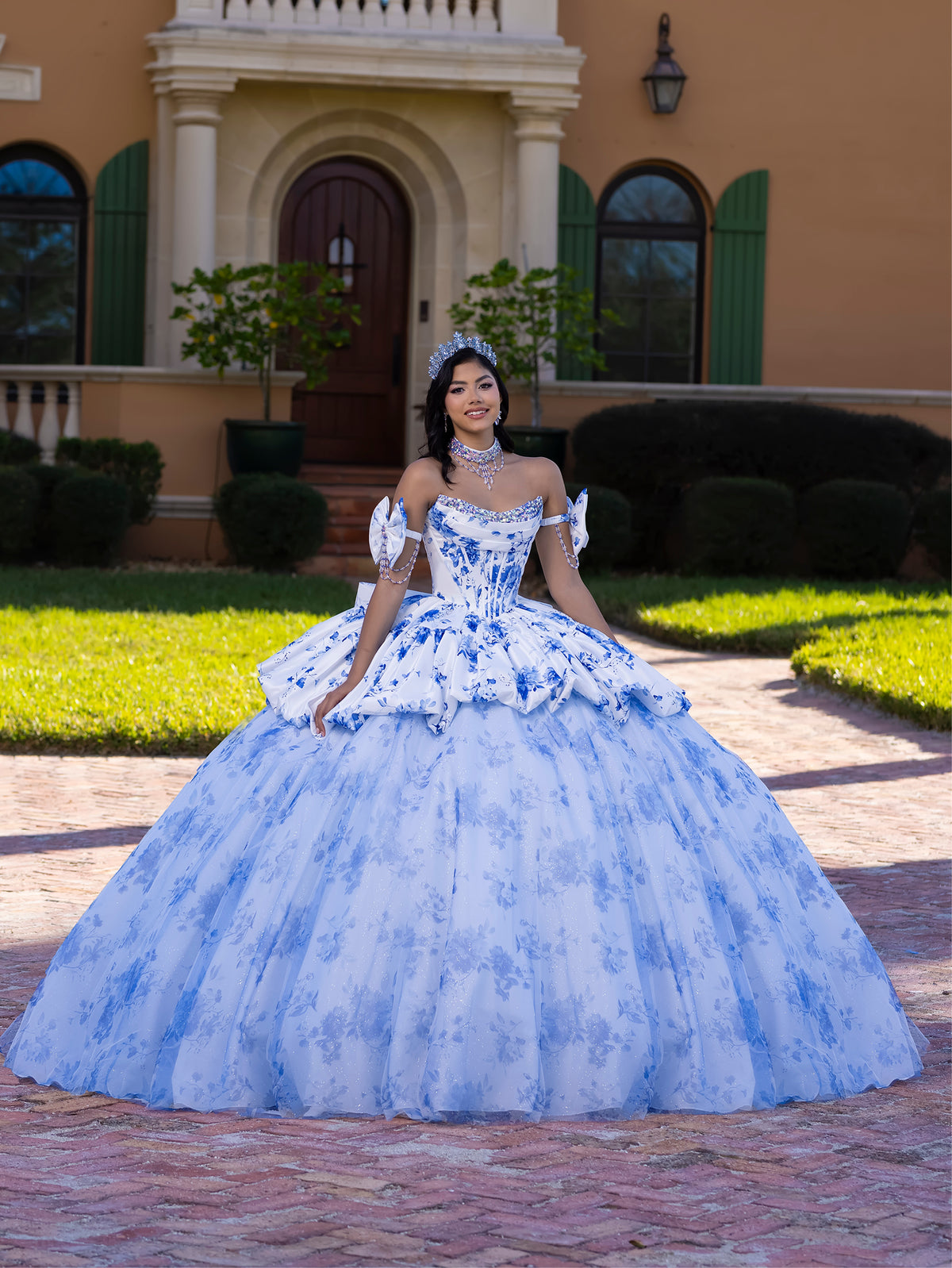 Woman in a blue floral dress standing in front of a house. / This gown features a full ball-gown skirt with delicate floral detailing and a fitted bodice with off-the-shoulder sleeves. A long, flowing train completes the elegant, fairytale look.