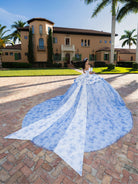 Woman in a blue gown with a long train standing in front of a large house. / This gown features a full ball-gown skirt with delicate floral detailing and a fitted bodice with off-the-shoulder sleeves. A long, flowing train completes the elegant, fairytale look.
