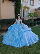 Woman in a blue quinceañera dress standing in front of a house. / This gown features an embellished off-the-shoulder bodice with layered textures and a full ball-gown skirt. The cascading layers create dramatic volume, while the back extends into a sweeping, embroidered train for an elegant, regal finish.