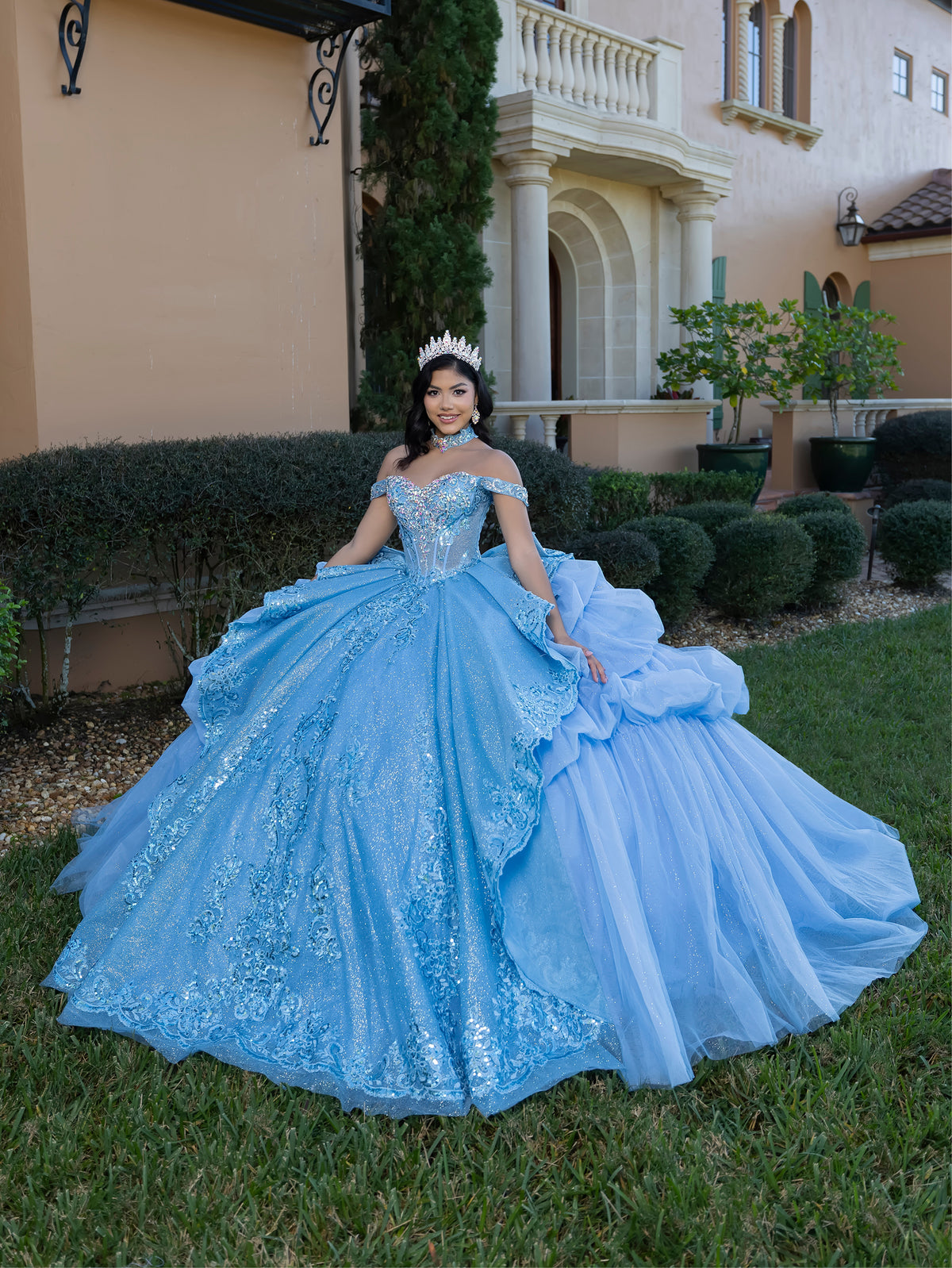 Woman in a blue quinceañera dress standing in front of a house. / This gown features an embellished off-the-shoulder bodice with layered textures and a full ball-gown skirt. The cascading layers create dramatic volume, while the back extends into a sweeping, embroidered train for an elegant, regal finish.