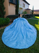 A woman photographed from behind wearing an elaborate light blue ball gown with a very large train, standing on a lawn in front of a large house. / This gown features an embellished off-the-shoulder bodice with layered textures and a full ball-gown skirt. The cascading layers create dramatic volume, while the back extends into a sweeping, embroidered train for an elegant, regal finish.