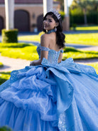 Woman in a blue formal gown with intricate details outdoors. / This gown features an embellished off-the-shoulder bodice with layered textures and a full ball-gown skirt. The cascading layers create dramatic volume, while the back extends into a sweeping, embroidered train for an elegant, regal finish.