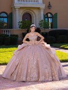 A woman wearing a voluminous champagne-beige ball gown with gold floral embroidery stands outdoors in front of a grand beige house with green shutters and manicured hedges. / This gown features an embellished off-the-shoulder bodice with layered textures and a full ball-gown skirt. The cascading layers create dramatic volume, while the back extends into a sweeping, embroidered train for an elegant, regal finish.