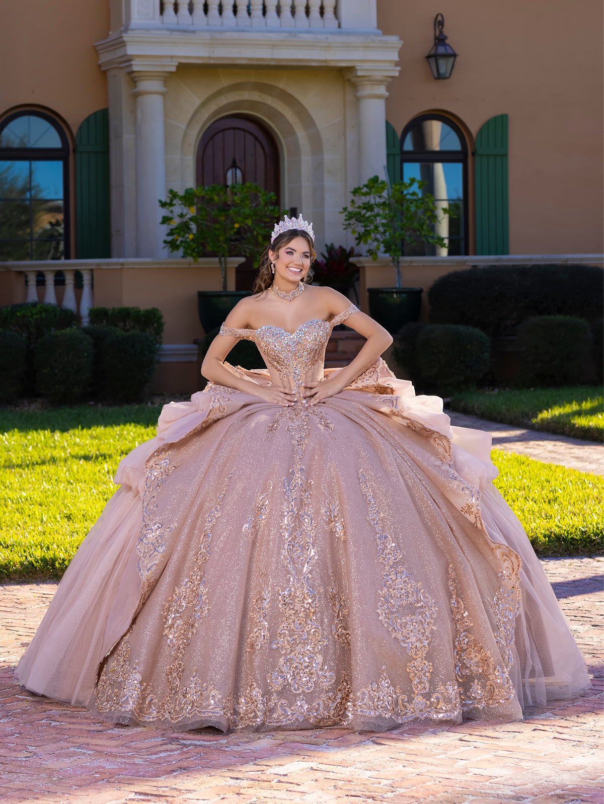 A woman wearing a voluminous champagne-beige ball gown with gold floral embroidery stands outdoors in front of a grand beige house with green shutters and manicured hedges. / This gown features an embellished off-the-shoulder bodice with layered textures and a full ball-gown skirt. The cascading layers create dramatic volume, while the back extends into a sweeping, embroidered train for an elegant, regal finish.