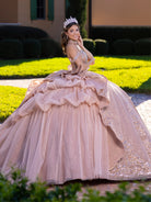 A woman wearing a voluminous blush pink ball gown with silver glitter and ruffled layers, standing outdoors in a garden setting with a matching tiara. / This gown features an embellished off-the-shoulder bodice with layered textures and a full ball-gown skirt. The cascading layers create dramatic volume, while the back extends into a sweeping, embroidered train for an elegant, regal finish.