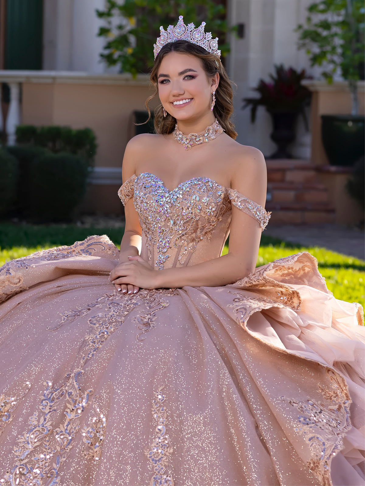 A young woman wearing an elaborate off-the-shoulder quinceañera gown in shimmering blush pink and silver, complete with a matching tiara and choker, posing outdoors in front of a house. / This gown features an embellished off-the-shoulder bodice with layered textures and a full ball-gown skirt. The cascading layers create dramatic volume, while the back extends into a sweeping, embroidered train for an elegant, regal finish.
