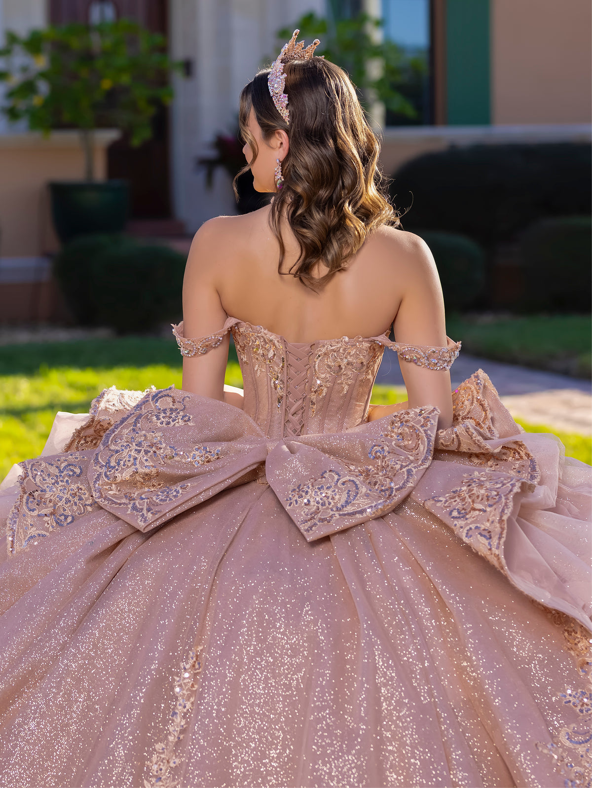 Woman in a sparkling pink gown with a large bow at the back, standing outdoors. / This gown features an embellished off-the-shoulder bodice with layered textures and a full ball-gown skirt. The cascading layers create dramatic volume, while the back extends into a sweeping, embroidered train for an elegant, regal finish.