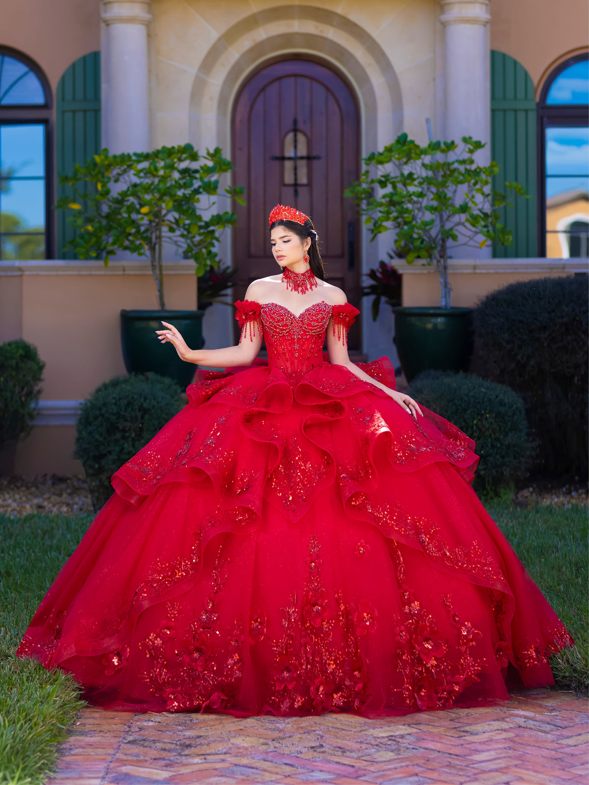 Woman in a red ball gown standing in front of a building with arched doorways. / This gown features an embellished off-the-shoulder bodice with delicate appliqué and a full tulle skirt decorated with floral embroidery. A structured accent at the waist and a long, ornate train complete the dramatic look.