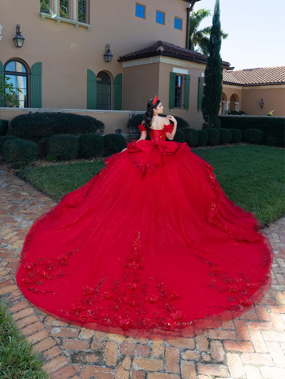 A woman wearing an elaborate bright red ball gown with a very large train stands outdoors in front of a beige house on a brick walkway. The dress has floral appliqué details and a fitted bodice. / This gown features an embellished off-the-shoulder bodice with delicate appliqué and a full tulle skirt decorated with floral embroidery. A structured accent at the waist and a long, ornate train complete the dramatic look.