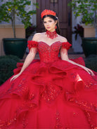 A woman wearing an elaborate off-the-shoulder red quinceañera ball gown with heavy beadwork and a matching red crown, posed outdoors in front of a house entrance. / This gown features an embellished off-the-shoulder bodice with delicate appliqué and a full tulle skirt decorated with floral embroidery. A structured accent at the waist and a long, ornate train complete the dramatic look.