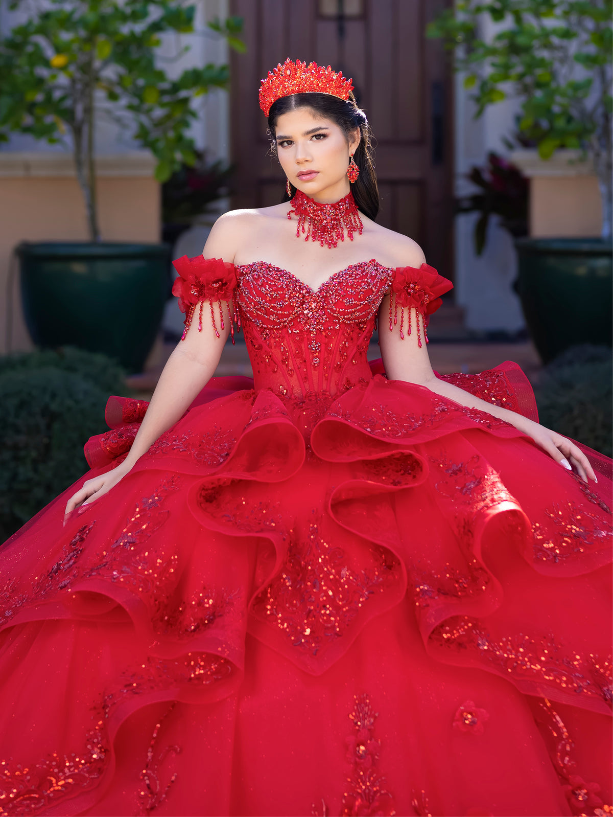 A woman wearing an elaborate off-the-shoulder red quinceañera ball gown with heavy beadwork and a matching red crown, posed outdoors in front of a house entrance. / This gown features an embellished off-the-shoulder bodice with delicate appliqué and a full tulle skirt decorated with floral embroidery. A structured accent at the waist and a long, ornate train complete the dramatic look.