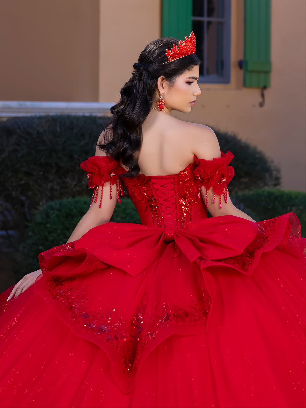 A woman wearing a bright red off-the-shoulder quinceañera dress with a large bow at the back, sequined details, and a matching red crown, photographed outdoors in front of a building with green shutters. / This gown features an embellished off-the-shoulder bodice with delicate appliqué and a full tulle skirt decorated with floral embroidery. A structured accent at the waist and a long, ornate train complete the dramatic look.