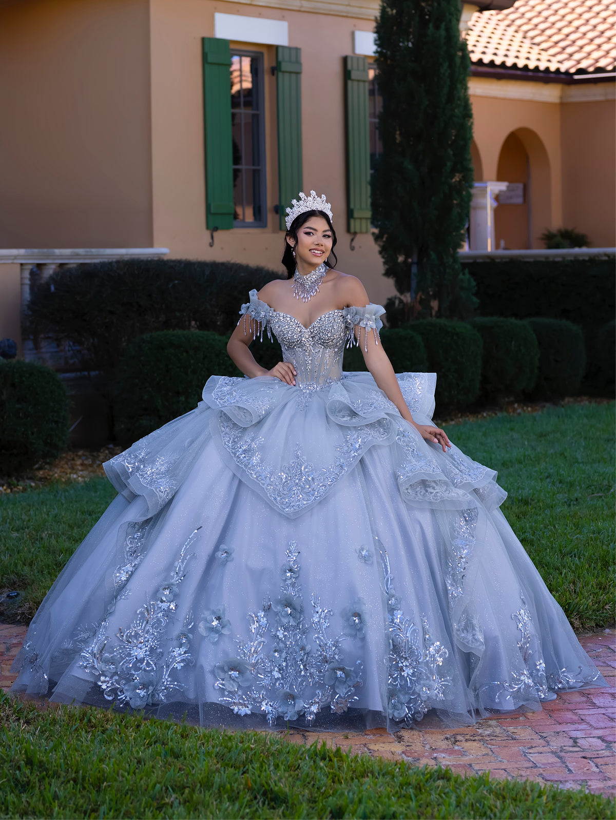 A woman wearing an elaborate light blue ball gown with silver floral embroidery and a matching tiara, standing outdoors on a brick path in front of a house with green shutters and trimmed hedges. / This gown features an embellished off-the-shoulder bodice with delicate appliqué and a full tulle skirt decorated with floral embroidery. A structured accent at the waist and a long, ornate train complete the dramatic look.