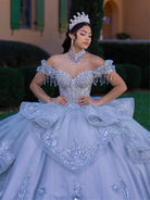 A woman wearing an elaborate off-the-shoulder quinceañera gown in icy light blue with heavy silver beadwork and sequins, matching tiara, and matching statement necklace, posing outdoors in front of a building and hedges. / This gown features an embellished off-the-shoulder bodice with delicate appliqué and a full tulle skirt decorated with floral embroidery. A structured accent at the waist and a long, ornate train complete the dramatic look.