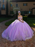 A woman wearing a voluminous lavender off-the-shoulder ball gown with silver floral embroidery and a matching tiara, standing on a brick path in front of a large villa. / This ball gown features an embellished off-the-shoulder bodice, and a full skirt decorated with intricate floral appliqué, finished with a back bow.