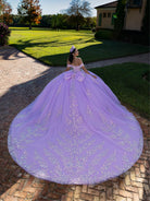 A woman wearing a voluminous lavender ball gown with silver floral embroidery stands on a brick path in a garden courtyard, showing the large train of the dress. / This ball gown features an embellished off-the-shoulder bodice, and a full skirt decorated with intricate floral appliqué, finished with a back bow.