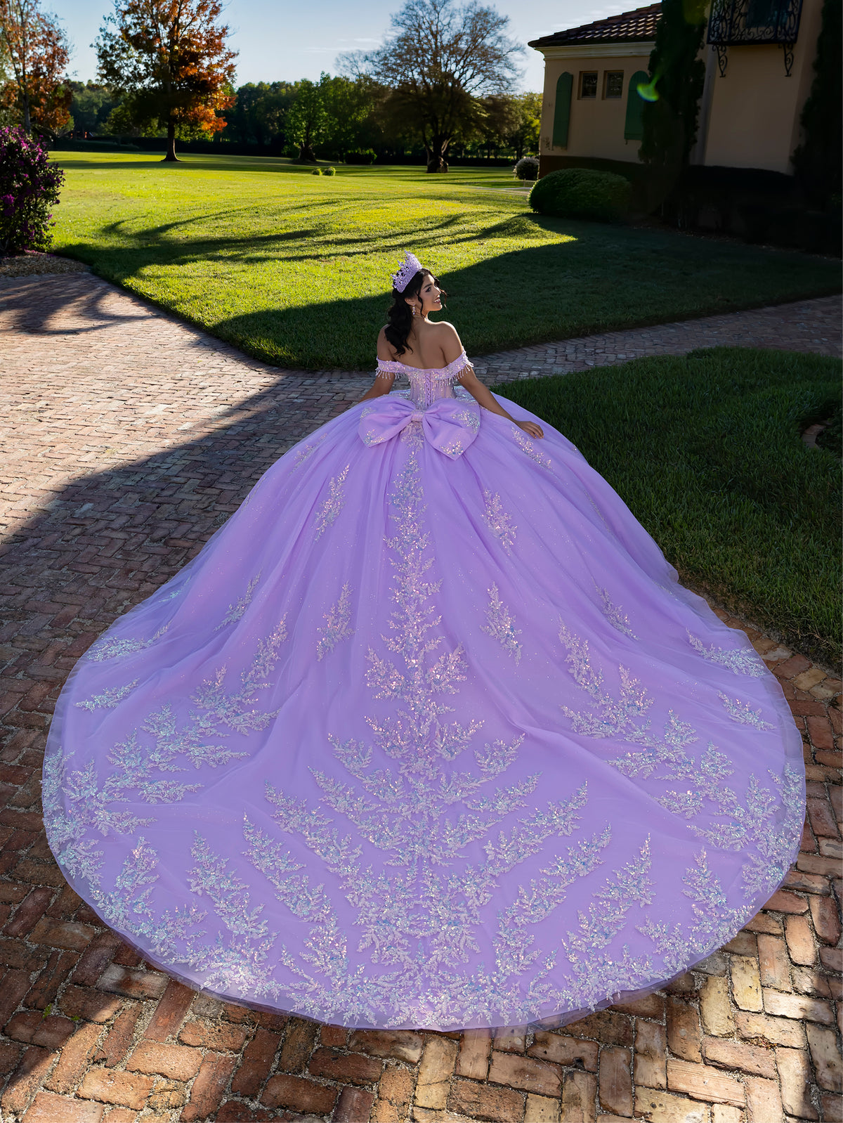 A woman wearing a voluminous lavender ball gown with silver floral embroidery stands on a brick path in a garden courtyard, showing the large train of the dress. / This ball gown features an embellished off-the-shoulder bodice, and a full skirt decorated with intricate floral appliqué, finished with a back bow.