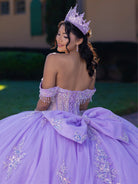 A woman wearing a lavender off-the-shoulder quinceañera ball gown with a large bow at the back, embellished with sparkling silver and blue beading, and wearing a matching lavender tiara, photographed outdoors. / This ball gown features an embellished off-the-shoulder bodice, and a full skirt decorated with intricate floral appliqué, finished with a back bow.