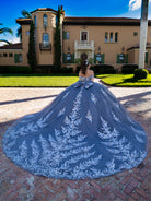 A woman stands outdoors in front of a large beige mansion on a brick patio, wearing a voluminous off-the-shoulder ball gown with a long train. The dress is dusty slate blue with intricate white floral embroidery and a large bow at the waist. / This ball gown features an embellished off-the-shoulder bodice, and a full skirt decorated with intricate floral appliqué, finished with a back bow.