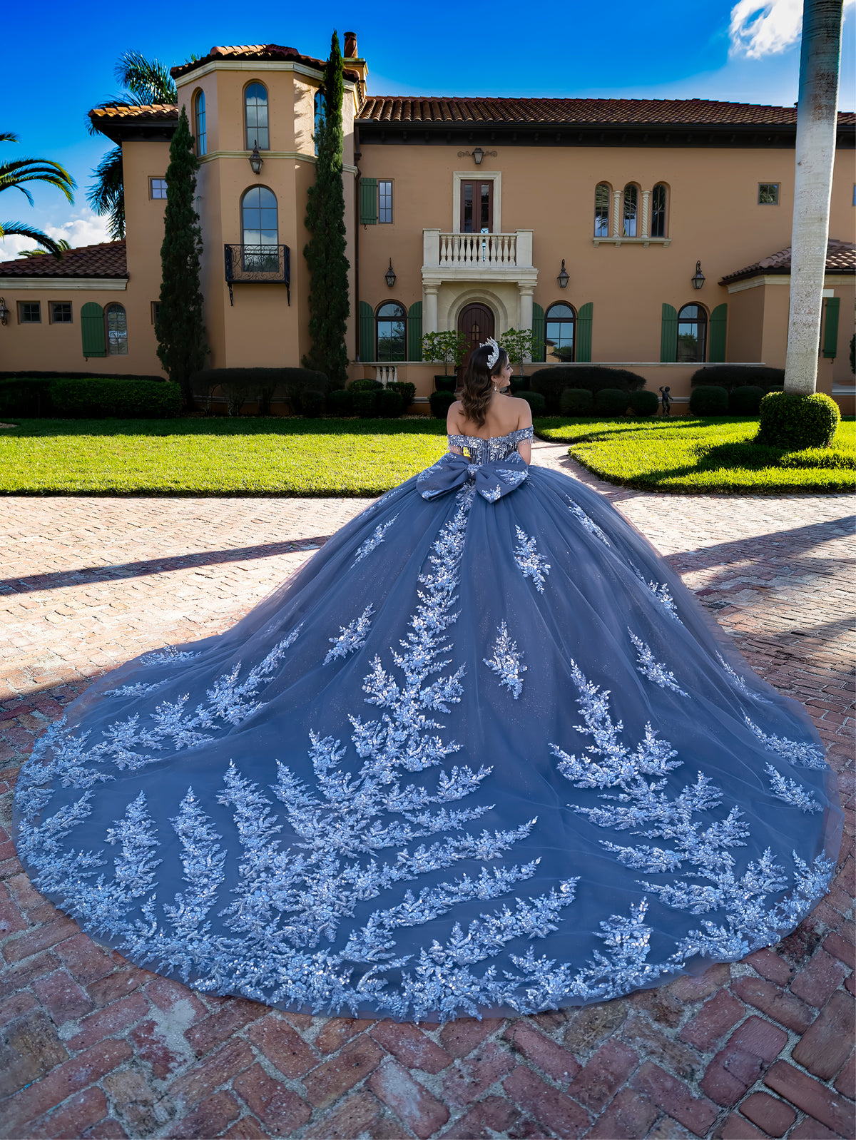 A woman stands outdoors in front of a large beige mansion on a brick patio, wearing a voluminous off-the-shoulder ball gown with a long train. The dress is dusty slate blue with intricate white floral embroidery and a large bow at the waist. / This ball gown features an embellished off-the-shoulder bodice, and a full skirt decorated with intricate floral appliqué, finished with a back bow.