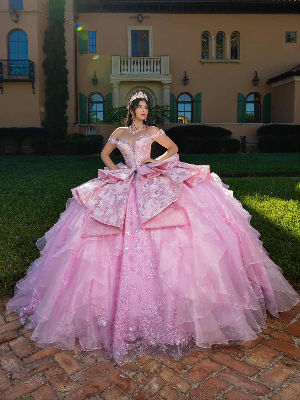 Woman in a pink gown standing in front of a mansion / This gown features an off-the-shoulder bodice with shimmering embroidery and a full, layered ball-gown skirt. Ruffled tiers and floral accents add volume, while a long, flowing train completes the elegant look.