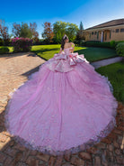 Pink ball gown with large bow on a person standing outdoors. / This gown features an off-the-shoulder bodice with shimmering embroidery and a full, layered ball-gown skirt. Ruffled tiers and floral accents add volume, while a long, flowing train completes the elegant look.