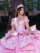 Woman in a pink gown with floral patterns and a tiara, sitting outdoors. / This gown features an off-the-shoulder bodice with shimmering embroidery and a full, layered ball-gown skirt. Ruffled tiers and floral accents add volume, while a long, flowing train completes the elegant look.