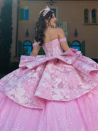 Woman in a pink ball gown with floral patterns and a tiara, standing in front of a building. / This gown features an off-the-shoulder bodice with shimmering embroidery and a full, layered ball-gown skirt. Ruffled tiers and floral accents add volume, while a long, flowing train completes the elegant look.