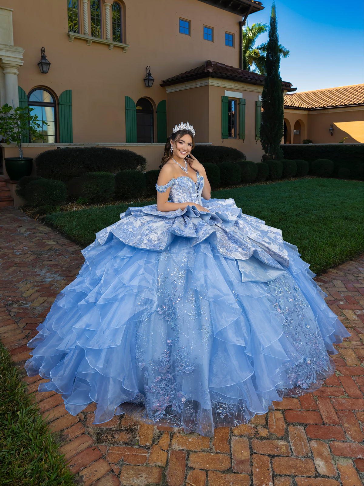 Woman in a blue formal gown standing on a brick path with a building in the background / This gown features an off-the-shoulder bodice with shimmering embroidery and a full, layered ball-gown skirt. Ruffled tiers and floral accents add volume, while a long, flowing train completes the elegant look.